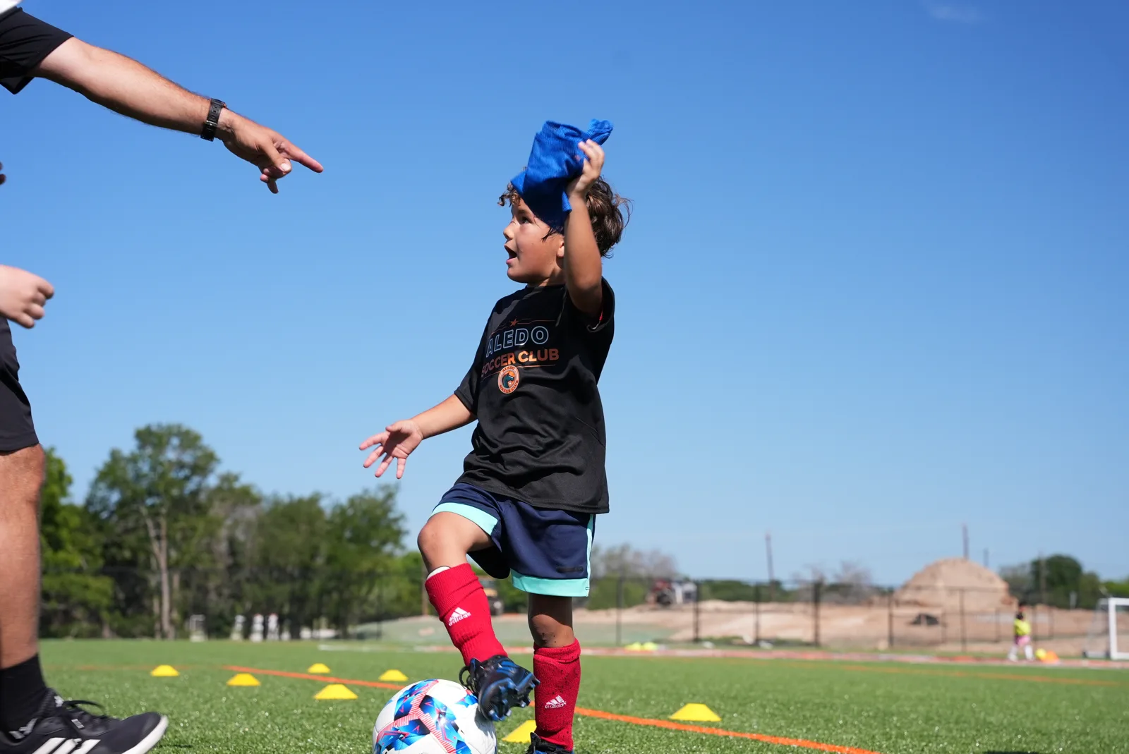 A young Little Aledos player in his Aledo Soccer Club shirt, ball at his feet, as his coach points out the next move at The Stable